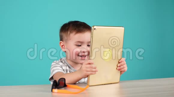 可爱的小男孩用平板电脑坐在桌子上孤立在蓝色上视频的预览图