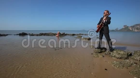 吉他手在沙滩上感情地演奏女孩坐在海边的岩石上视频的预览图