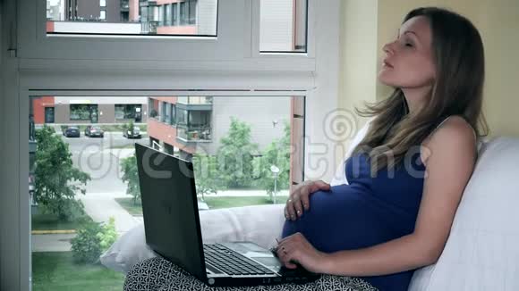 疲惫的自由职业孕妇坐在窗户旁手提电脑工作视频的预览图
