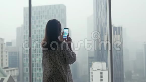 吉隆坡一名女游客站在窗口用手机拍摄摩天大楼高度视频的预览图