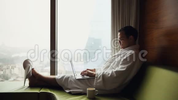 年轻的微笑商人在手提电脑上工作穿着白色浴袍坐在窗边喝杯咖啡视频的预览图