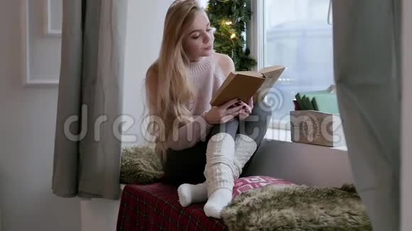 年轻的金发女人穿着紧身裤坐在窗户上读一本书圣诞节内部视频的预览图