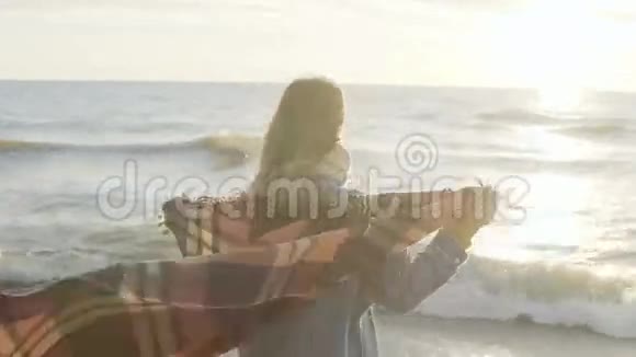 自由年轻女子站在海边用格子布举起手在寒冷的风日快乐的女性在海滩上视频的预览图