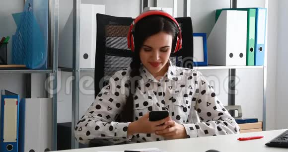 女商人戴着红色耳机在现代办公室里使用智能手机视频的预览图