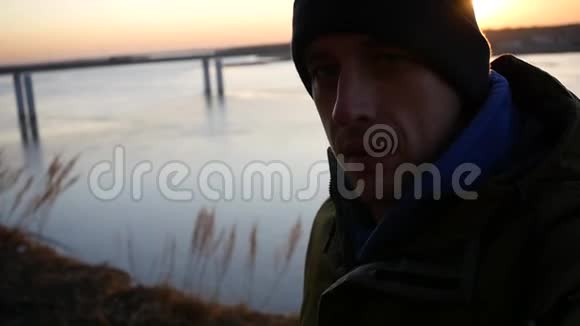 一个戴着帽子的年轻人在河边的一座桥上带着夕阳做自拍看着镜头慢动作1920x1080视频的预览图