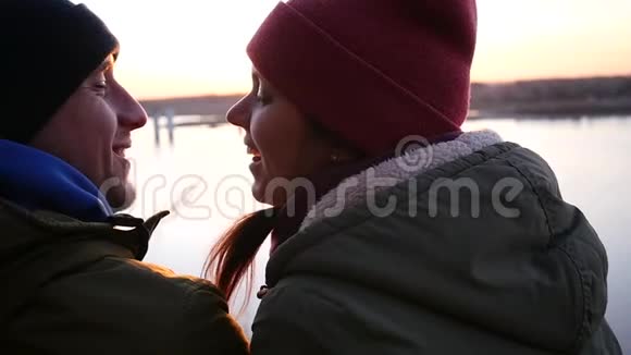 爱和幸福的一对夫妇坐在河岸上在夕阳的背景下互相微笑慢动作视频的预览图