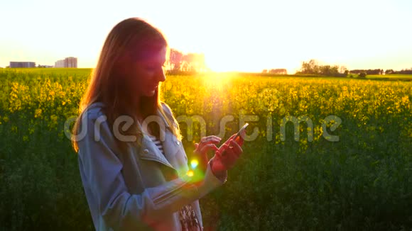 美丽的女人在夕阳下在黄花的田野里用电话视频的预览图