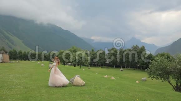 红发新娘穿着奢华的礼服在高山的映衬下沿着绿草地散步婚礼日视频的预览图