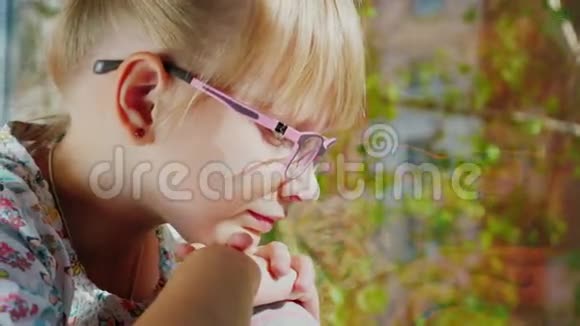 金发女孩6岁戴着粉红色眼镜看着窗外窗外春天听到一棵树的绿叶视频的预览图