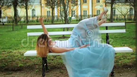 漂亮的女孩穿着白色和蓝色的连衣裙躺在长凳上腿穿着高跟鞋在长凳上休息她带着视频的预览图