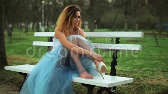 穿着白色和蓝色连衣裙的漂亮女孩坐在Parkway的长凳上穿上高跟鞋扣鞋在公园里交谈视频的预览图