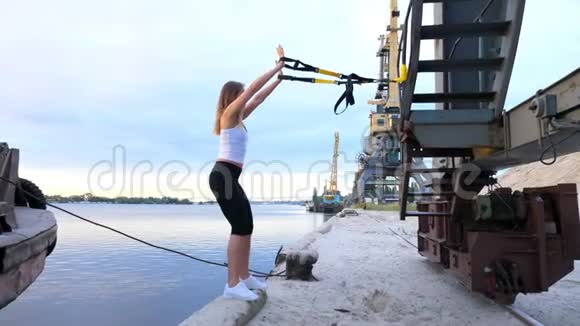 美丽的女士在悬吊带TRX的帮助下锻炼她的肌肉在背景上可以看到河流货物视频的预览图