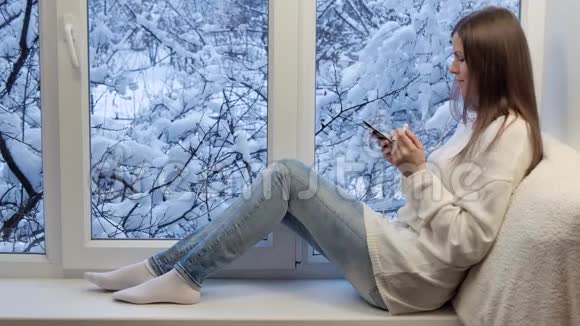 漂亮的女孩坐在窗台上看着窗户用智能手机冬天在外面视频的预览图