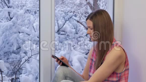 漂亮的女孩坐在窗台上看着窗户用智能手机冬天在外面视频的预览图