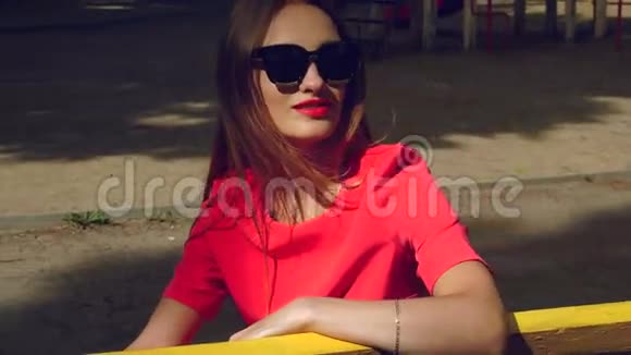 穿着红色西装戴着黑色太阳镜的快乐迷人女孩坐在公园的长凳上视频的预览图