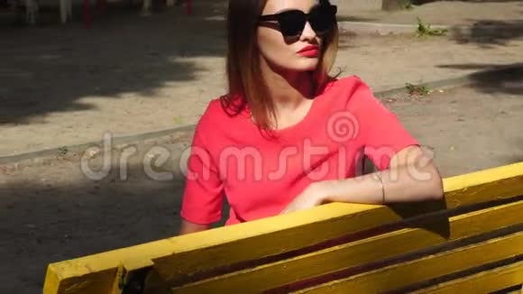 漂亮的女士戴着太阳镜穿着红色西装坐在公园的黄色长凳上视频的预览图