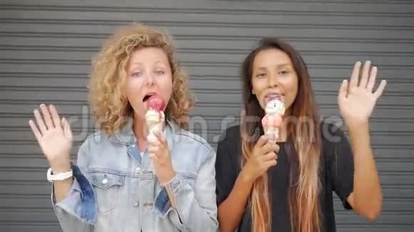 两个年轻的快乐无职业混合种族女孩挥舞手在相机和吃冰淇淋锥滑鼠生活高清视频的预览图