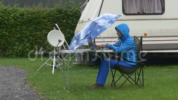 一个旅行者在蓝伞下的笔记本电脑下工作在大篷车附近下雨视频的预览图