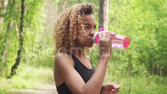 黑色女人从粉红色的瓶子里喝黑人年轻女子慢跑休息的肖像视频的预览图