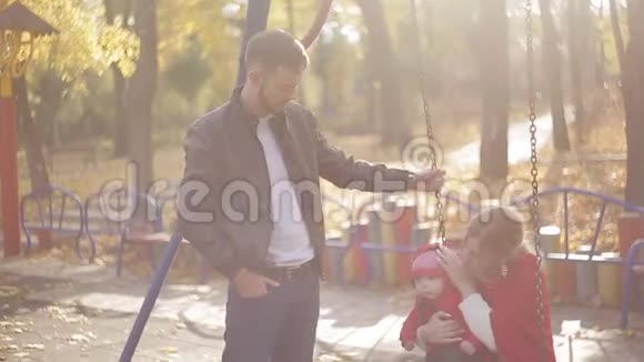 年轻的爸爸妈妈和一个婴儿在操场上玩耍公园里为孩子们准备的秋千和旋转木马视频的预览图