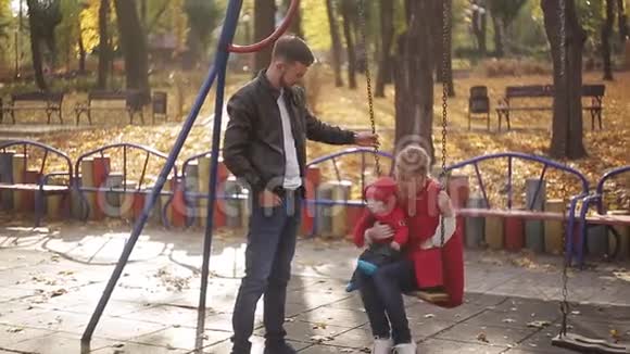 年轻的爸爸妈妈和一个婴儿在操场上玩耍公园里为孩子们准备的秋千和旋转木马视频的预览图
