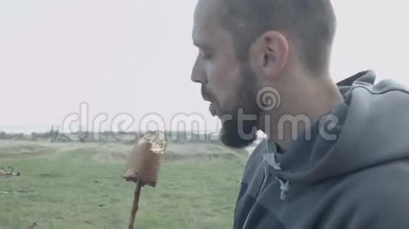 一个秃头英俊的人在大自然中吃烤面包烤在火上的棍子上视频的预览图