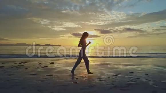 黎明时分女孩的剪影慢慢地沿着潮湿的海滩漫步视频的预览图