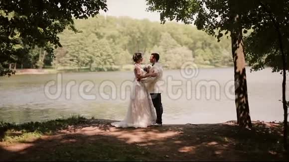 婚礼当天的恋人们站在河岸上互相看着对方接吻穿着漂亮裙子的快乐新娘视频的预览图