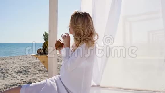女孩在海滩上休息喝椰子汁美丽的女人坐在平房里岸上有白色的窗帘视频的预览图