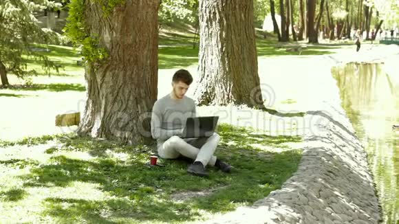 一个年轻人坐在树荫下的小溪边手里拿着咖啡杯和笔记本电脑视频的预览图