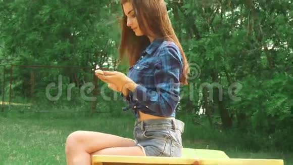 年轻女孩坐在公园的长凳上用手机视频的预览图