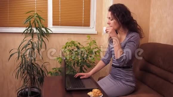 有笔记本电脑和咖啡的女孩一个穿灰色连衣裙的女人在笔记本电脑后面的室内喝咖啡在沙发上喝咖啡的年轻女子视频的预览图