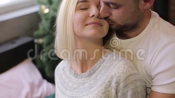 年轻夫妇坐在圣诞树背景上的床上在家过喜庆的夜晚男人亲吻女人视频的预览图
