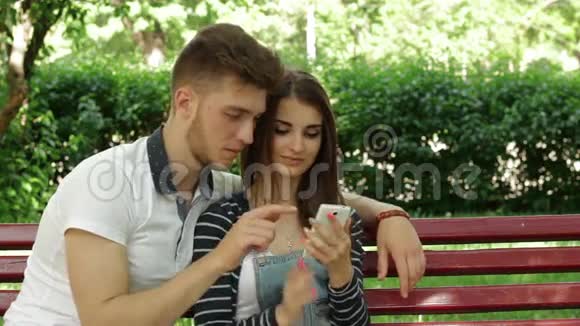 男人和女人坐在长凳上在电话里看照片视频的预览图