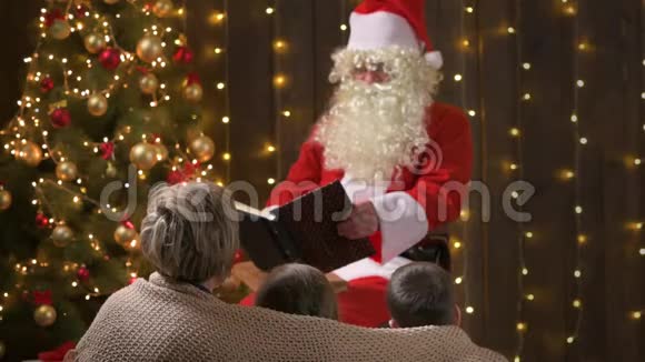 圣诞老人为家人读书母亲和孩子们坐在室内靠近装饰好的圣诞树和灯圣诞快乐视频的预览图