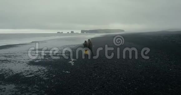 在冰岛黑色火山海滩散步的年轻时尚夫妇的空中景观男人和女人从波浪中奔跑视频的预览图