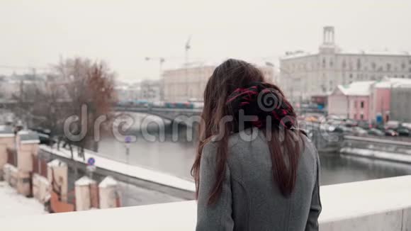 后景站在桥上悲伤的黑发女孩叹了口气看着白雪覆盖的冬季小镇视频的预览图