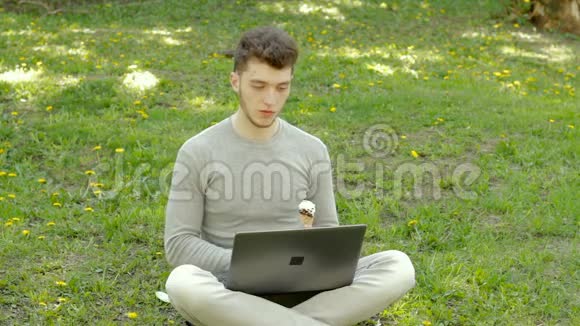在草地上的公园里一个学生用笔记本电脑工作吃一个冰淇淋筒视频的预览图