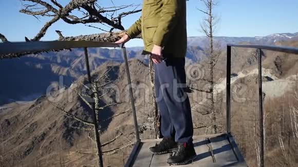 一个年轻人站在一棵老干树附近的山顶上看着山谷和山脉的全景视频的预览图