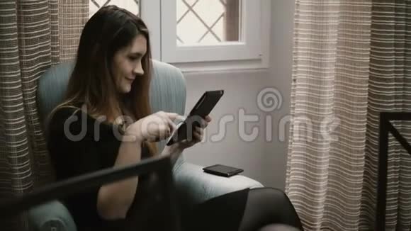 年轻的美女坐在家里的椅子上使用智能技术在平板电脑上工作视频的预览图