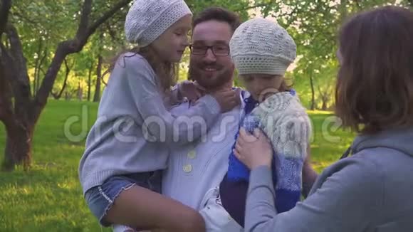 幸福的家庭拥抱在草地上父母和他们的两个女儿正在公园草坪上拥抱开着花的果园视频的预览图