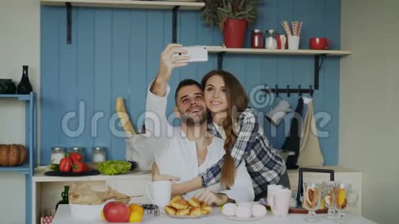 年轻的幸福夫妇在家里的厨房里吃早餐时正在自拍视频的预览图