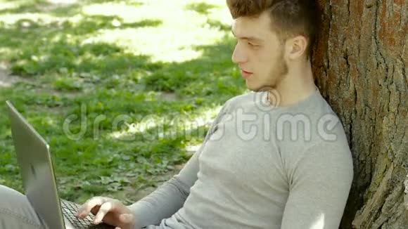 年轻漂亮的学生在公园的一棵树下的笔记本电脑上写字视频的预览图
