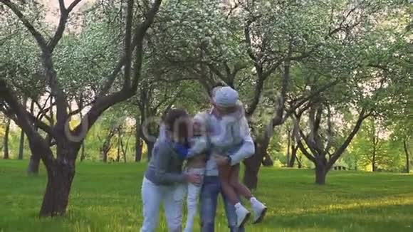 幸福的家庭拥抱在草地上父母和他们的两个女儿正在公园草坪上拥抱开着花的果园视频的预览图