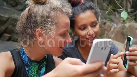 热带森林中使用移动电话坐瀑布的年轻徒步旅行者女孩高清慢速运动视频的预览图
