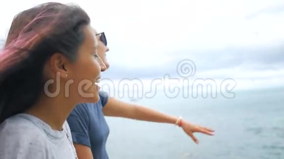 两个年轻的无忧无虑的时髦女孩坐在岩石上张开双臂欣赏海景高清慢动作关闭苏视频的预览图