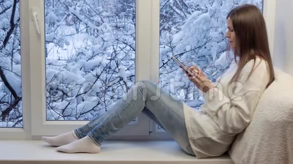 漂亮的女孩坐在窗台上用带耳机的平板电脑冬天在外面视频的预览图