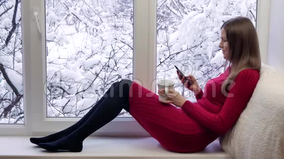 穿着红裙子的漂亮女孩坐在窗台上喝茶用智能手机冬天在外面视频的预览图