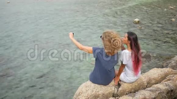 两个年轻的无顾客少女用手机拍自拍坐在海边的岩石上高清慢速运动视频的预览图