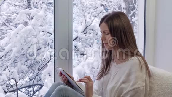 漂亮的女孩坐在窗台上用带耳机的平板电脑冬天在外面视频的预览图